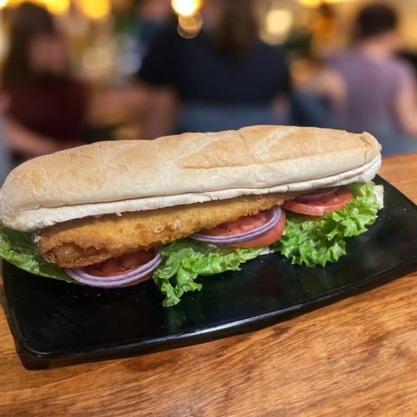 A fried fish sandwich on a baguette with lettuce, tomato, and red onion on a black plate.