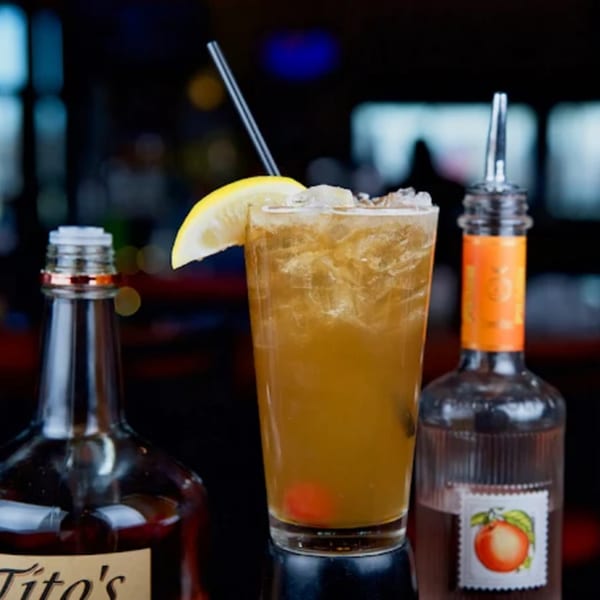 A tall glass of iced tea with a lemon slice and cherry, flanked by liquor bottles.
